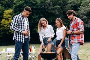 ragazzi in piedi accanto a un barbecue. ragazze che cucinano e uomini che tengono una birra e sorridono foto