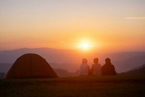gruppo di amici seduta di il tenda durante durante la notte campeggio mentre guardare a il bellissimo Visualizza punto tramonto al di sopra di il montagna per all'aperto avventura vacanza viaggio foto