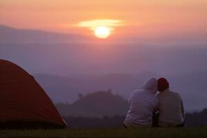 coppia è seduta di il tenda durante durante la notte campeggio mentre guardare a il bellissimo panoramico tramonto al di sopra di il montagna per all'aperto avventura vacanza viaggio foto