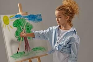 affascinante scuola ragazza è pittura con un' acquerello spazzola su un cavalletto, in piedi su un' grigio sfondo. foto