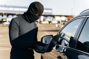 mezzo busto ritratto del ladro d'auto con il sistema di allarme hacking del computer portatile mentre si trova vicino all'auto durante il giorno. foto d'archivio