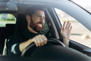 bel giovane autista sorridente di auto agitando la mano in segno di saluto mentre si guida l'auto con emozioni di piacere. concetto di trasporto foto