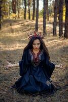 bellissimo strega nel Nero, lungo vestire, con rosso corona nel sua lungo capelli. in posa seduta nel pino foresta. incantesimi, Magia e stregoneria. pieno lunghezza. foto