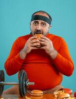 di mezza età uomo con barba, vestito nel un' rosso dolcevita, fascia per capelli, in posa con hamburger e francese patatine fritte. blu sfondo. avvicinamento. veloce cibo. foto