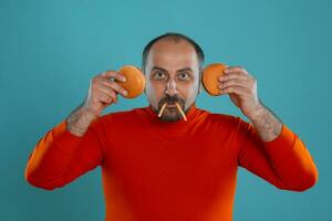 avvicinamento ritratto di un' di mezza età uomo con barba, vestito nel un' rosso dolcevita, in posa con hamburger contro un' blu sfondo. veloce cibo. foto