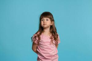 bellissimo poco ragazza indossare nel un' rosa maglietta è in posa contro un' blu studio sfondo. foto