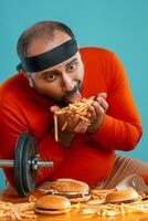 di mezza età uomo con barba, vestito nel un' rosso dolcevita, fascia per capelli, in posa con hamburger e francese patatine fritte. blu sfondo. avvicinamento. veloce cibo. foto