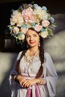 brunetta ragazza nel un' bianca ucraino autentico nazionale costume e un' ghirlanda di fiori è in posa contro un' bianca capanna. avvicinamento. foto