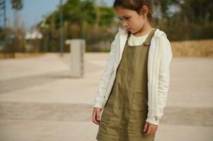 ritratto di un' bambino ragazza nel cachi vestito e beige felpa, guardare fuori uso, esprimendo tristezza in piedi su urbano sfondo foto