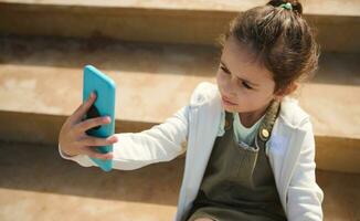 adorabile elementare età caucasico bambino ragazza Tenere mobile Telefono e fabbricazione autoscatto all'aperto foto