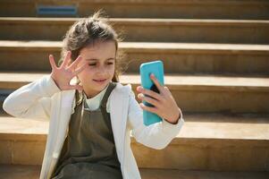 caucasico ragazza utilizzando smartphone, in linea chattare, comunicazione, sorridente agitando Ciao con mano, parlando attraverso video collegamento, foto