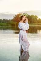 bellissimo nero dai capelli ragazza nel bianca Vintage ▾ vestito e ghirlanda di fiori in piedi nel acqua di lago. sole bagliore. foto