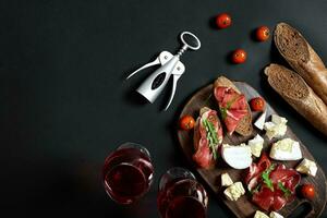 prosciutto, salame, baguette fette, pomodori e pazzo rustico di legno asse, Due bicchieri di rosso vino al di sopra di nero sfondo foto