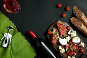 delizioso aperitivo per vino - prosciutto, formaggio, baguette fette, pomodori, servito su un' di legno asse, e bicchiere con rosso vino su nero superficie foto