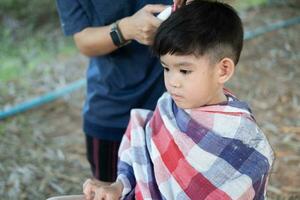 barbiere taglio capelli di un asiatico ragazzo nel un Aperto spazio pieno con alberi. foto