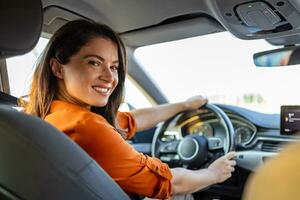 giovane donna seduta su un' autisti posto a sedere nel il auto e guardare a telecamera al di sopra di il spalla. ritratto di piacevole femmina con positivo espressione, essere soddisfatto con indimenticabile viaggio di macchina. foto