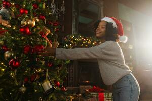 allegro Natale. africano americano donna decorazione Natale albero. contento ragazza vicino tradizionale Natale albero con classico rosso d'oro decorazioni ornamento, Natale vigilia a casa tempo per celebrazione foto