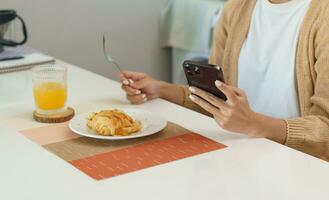 contento bellissimo asiatico donna con Telefono mangiare fresco insalata a tavolo nel cucina e utilizzando applicazione sms su smartphone mentre mangiare salutare cibo. foto