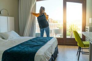 turista donna nel il Hotel Camera da letto con sua bagaglio in piedi vicino il finestra. viaggio e vacanza concetto. foto