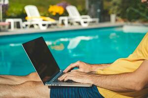 giovane uomo Lavorando con il computer portatile Il prossimo per il piscina durante un' soleggiato giorno. attraente uomo Lavorando. libero professionista. viaggiatore. foto