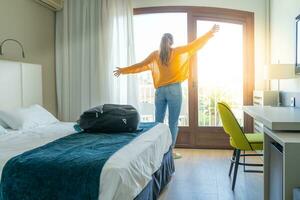 turista donna nel il Hotel Camera da letto con sua bagaglio sollevato mani su e in piedi vicino il finestra. viaggio e vacanza concetto. foto