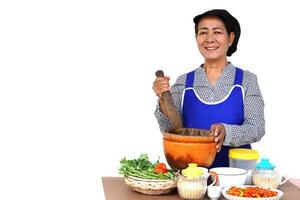 contento asiatico anziano donna è cucinando, indossa berretto, blu grembiule, detiene pestello e di legno mortaio. bianca sfondo. concetto, cucinando per famiglia. tailandese cucina stile di vita. anziano attività. foto