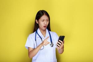 ritratto di un' bellissimo giovane donna nel un' giallo sfondo, asiatico donna pose con un' cellula Telefono mentre indossare un' medico uniforme e un' stetoscopio. foto