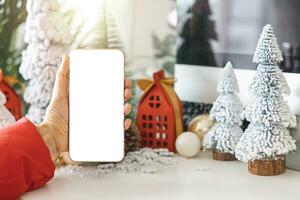 dolce mani culla un' smartphone con un' bianca schermo finto su di festivo scena un' bellissimo Natale albero adornato, con Natale palle, pino coni, e rosso Casa regalo scatola legato d'oro nastro. foto
