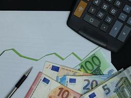 banconote europee, penna e calcolatrice su sfondo con linea verde di tendenza in aumento, vista dall'alto foto
