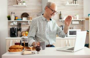 quotidiano vita di anziano uomo nel cucina durante prima colazione utilizzando il computer portatile Tenere un' tazza di caffè, parlando in linea con nipoti agitando a webcam. pensionato persona Lavorando a partire dal casa su virtuale incontro, conferenza foto