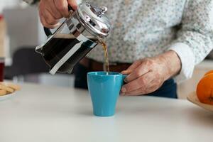 vicino su di anziano uomo mani scrosciante caffè nel tazza durante prima colazione seduta nel moderno cucina. anziano persona nel il mattina godendo fresco Marrone caffè espresso a partire dal Vintage ▾ tazza, filtro rilassare rinfresco foto