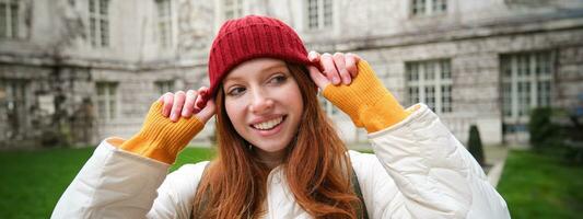 ritratto di sorridente testa Rossa donna mette su rosso cappello e sorrisi, indossare caldo Abiti mentre esplorando città strade foto