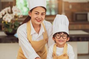 ritratto carino piccolo ragazzo asiatico chef con occhiali e sua madre che guarda alla macchina fotografica in cucina casalinga cucina felicemente e divertente. stili di vita delle persone e della famiglia. concetto di produzione di ingredienti alimentari fatti in casa foto