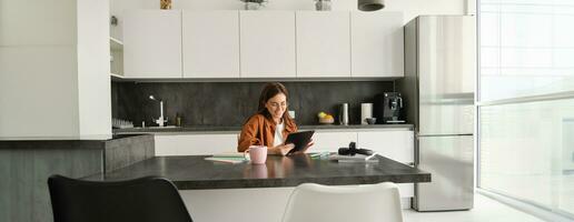 ritratto di giovane donna, alunno seduta nel cucina con tavoletta e taccuino, lettura notizia in linea, studiando a distanza, elearning a casa foto