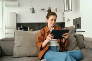 ritratto di giovane donna seduta su divano nel vivente camera, Guardando film su digitale tavoletta, lettura su dispositivo, godendo fine settimana a casa foto