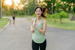 in forma asiatico giovane donna jogging nel parco sorridente contento in esecuzione e godendo un' salutare all'aperto stile di vita. femmina pareggiatore. fitness corridore ragazza nel pubblico parco. salutare stile di vita e benessere essere concetto foto