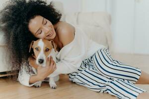 gioioso donna nel un' bianca maglione abbracciare sua cane, condivisione un' tenero momento a casa foto