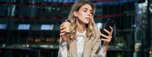 elegante aziendale donna nel completo da uomo, bevande sua caffè porta via e usi mobile Telefono App foto