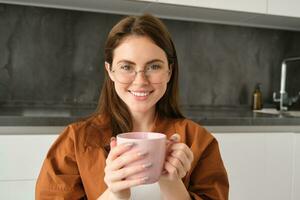 ritratto di bellissimo giovane donna nel occhiali, sorridente, riposo a casa, Tenere boccale con caffè, potabile tè foto