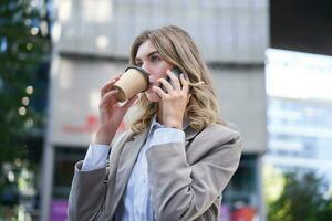 donna d'affari a piedi nel città centro, potabile caffè e parlando su mobile Telefono foto
