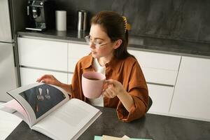 ritratto di bellissimo giovane donna nel cucina, seduta con un' prenotare, capovolgimento pagine, lettura e potabile caffè foto