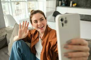 amichevole sorridente giovane donna, seduta su divano e agitando mano a smartphone telecamera, video chat, parlando su mobile Telefono, si connette per in linea conversazione su applicazione foto