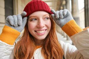 ritratto di giovane testa Rossa donna nel a maglia cappello e guanti, sorrisi e sembra a parte, passeggiate in giro città nel inverno foto