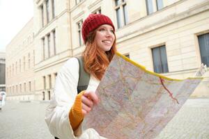bellissimo testa Rossa donna, turista con città carta geografica, esplora giro turistico storico punto di riferimento, a piedi in giro vecchio cittadina, sorridente felicemente foto