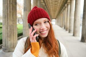 sorridente carino testa Rossa donna fa un' Telefono chiamata, detiene telefono vicino anno, ha mobile conversazione, utilizzando smartphone su strada foto