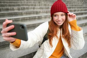 elegante giovane ragazza nel rosso cappello, prende fotografie su smartphone telecamera, fa autoscatto come lei si siede su le scale vicino Museo, in posa per foto con App filtro