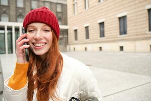 elegante moderno testa Rossa ragazza parla su mobile Telefono, fa un' telefono chiamata, chiamata qualcuno su smartphone App a partire dal fuori, sta su strada e sorrisi foto