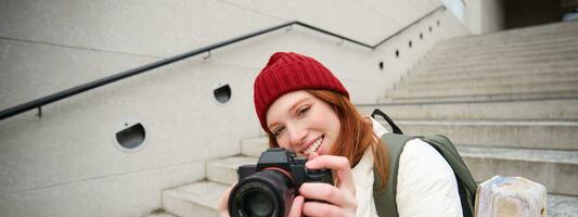 urbano persone e stile di vita. contento testa Rossa donna prende fotografie, Tenere professionale digitale telecamera, Fotografare su strade foto