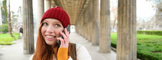 sorridente carino testa Rossa donna fa un' Telefono chiamata, detiene telefono vicino anno, ha mobile conversazione, utilizzando smartphone su strada foto