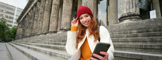 ritratto di giovane testa Rossa donna nel rosso cappello, seduta su scale, turista sembra a sua mobile Telefono, riposa su scala di Museo, si connette pubblico Wi-Fi foto